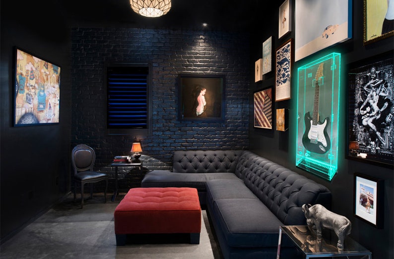 May include: A modern, dark-themed room with a black brick wall, a gray tufted sectional sofa, and a red ottoman. A guitar is displayed in a lighted acrylic case. Artwork and a small table with a lamp complete the space.