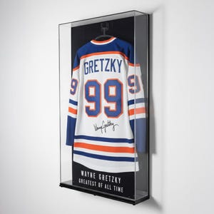 Puede incluir: Una camiseta de hockey enmarcada con el nombre "GRETZKY" y el n&uacute;mero "99" en la espalda. La camiseta es blanca con detalles azules y naranjas. El marco incluye el texto "WAYNE GRETZKY GREATEST OF ALL TIME".