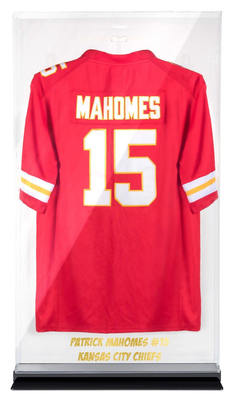 Puede incluir: Camiseta roja de los Kansas City Chiefs con el nombre "MAHOMES" y el n&uacute;mero 15 en blanco. La camiseta se exhibe en una vitrina. Las mangas tienen rayas blancas y amarillas. Texto "PATRICK MAHOMES #15 KANSAS CITY CHIEFS" en la parte inferior.