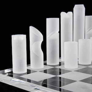 May include: A modern chess set with frosted glass pieces on a clear checkered board. The pieces are cylindrical with unique geometric designs, set against a black background. The set includes pawns, rooks, bishops, a queen, and a king.