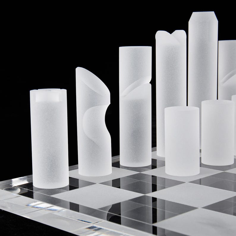 May include: A modern chess set with frosted glass pieces on a clear checkered board. The pieces are cylindrical with unique geometric designs, set against a black background. The set includes pawns, rooks, bishops, a queen, and a king.