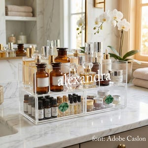 Op de afbeelding: Een doorzichtige acryl organizer gevuld met diverse parfumflesjes, huidverzorgingsproducten en etherische oliën. De organizer heeft twee niveaus en is gelabeld met de naam "alexandra" in witte letters. De achtergrond omvat een marmeren aanrecht en een witte orchidee.