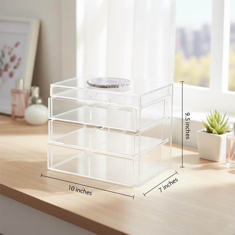 K&ouml;nnte beinhalten: Ein klarer Acryl-Organizer mit drei Schubladen, 25,4 cm breit, 17,8 cm tief und 24,1 cm hoch. Obenauf liegt ein dekorativer lila-wei&szlig;er Stein. Der Organizer steht auf einer hellen Holzoberfl&auml;che.