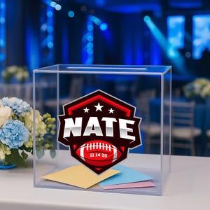 May include: A clear acrylic box with a slot on top, featuring a custom logo with the name "NATE" and a football graphic. The logo is red, white, and black. Inside the box are several colorful envelopes. The background is blurred.