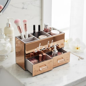 May include: A rose gold mirrored makeup organizer with the name "stella" in white script. The organizer has multiple compartments for brushes, lipsticks, and other cosmetics, and two drawers with crystal-like handles. The organizer is on a white marble surface.