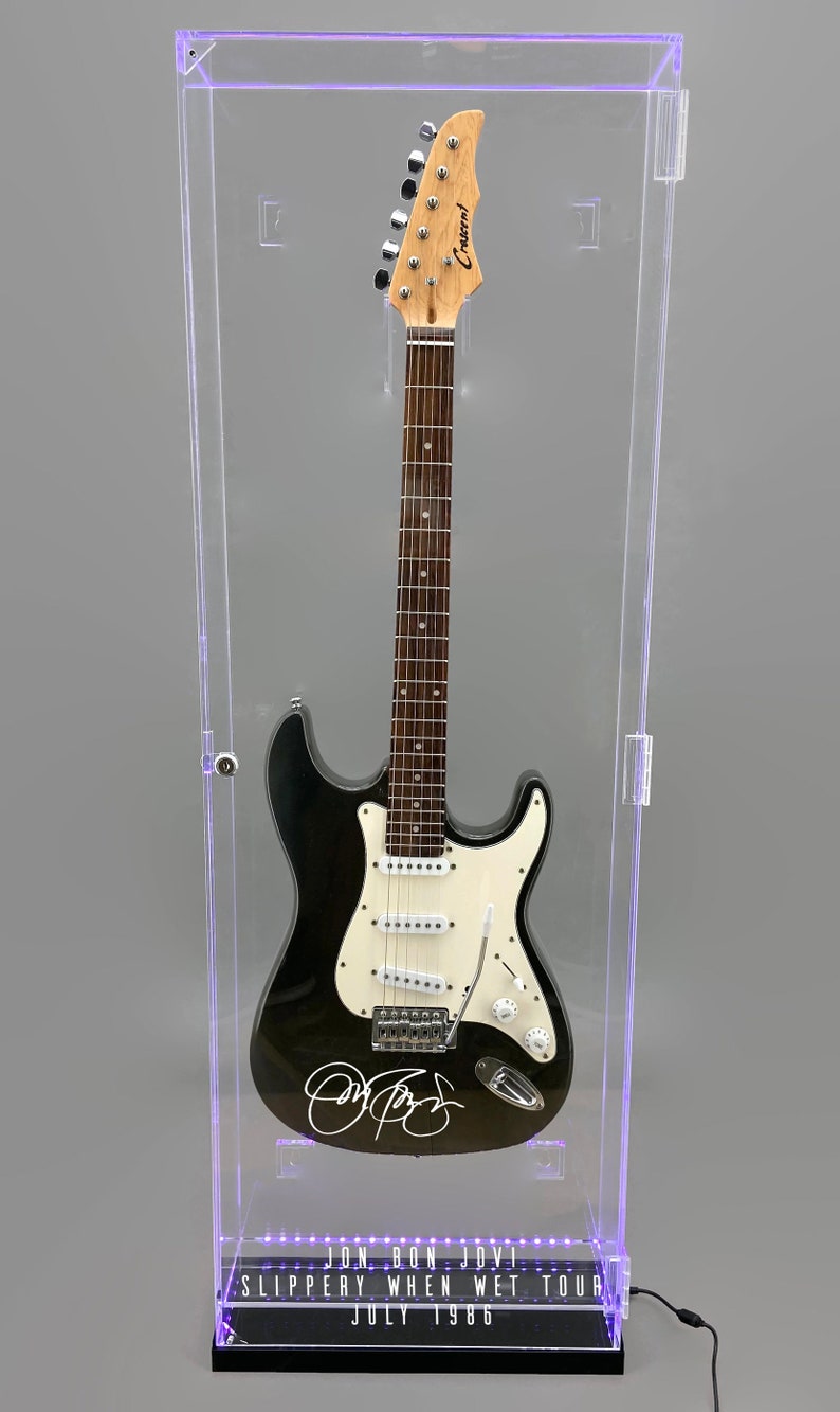 May include: Black and white electric guitar in a clear display case. The guitar has a signature on the body and the text "JON BON JOVI SLIPPERY WHEN WET TOUR JULY 1986" at the base. The case has a black base and a light.