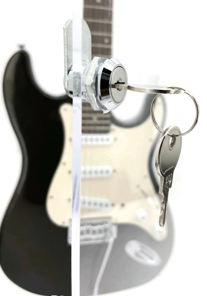 May include: A chrome-finished lock with a key inserted, set against a blurred background of a black and white electric guitar. The key is on a ring with another key.