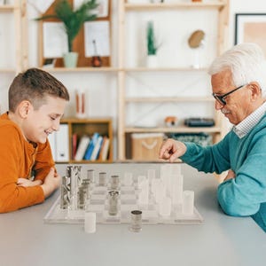May include: A clear acrylic chess set with silver and white pieces is set up on a gray table. A young person in an orange shirt and an older person in a blue sweater are playing chess. The chess board is transparent.