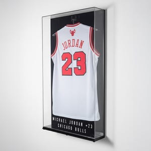 Puede incluir: Una camiseta de baloncesto blanca con detalles rojos y el n&uacute;mero 23, exhibida en una vitrina con marco negro. La camiseta tiene la palabra "JORDAN" en el pecho. La vitrina incluye el texto "MICHAEL JORDAN - 23 CHICAGO BULLS".