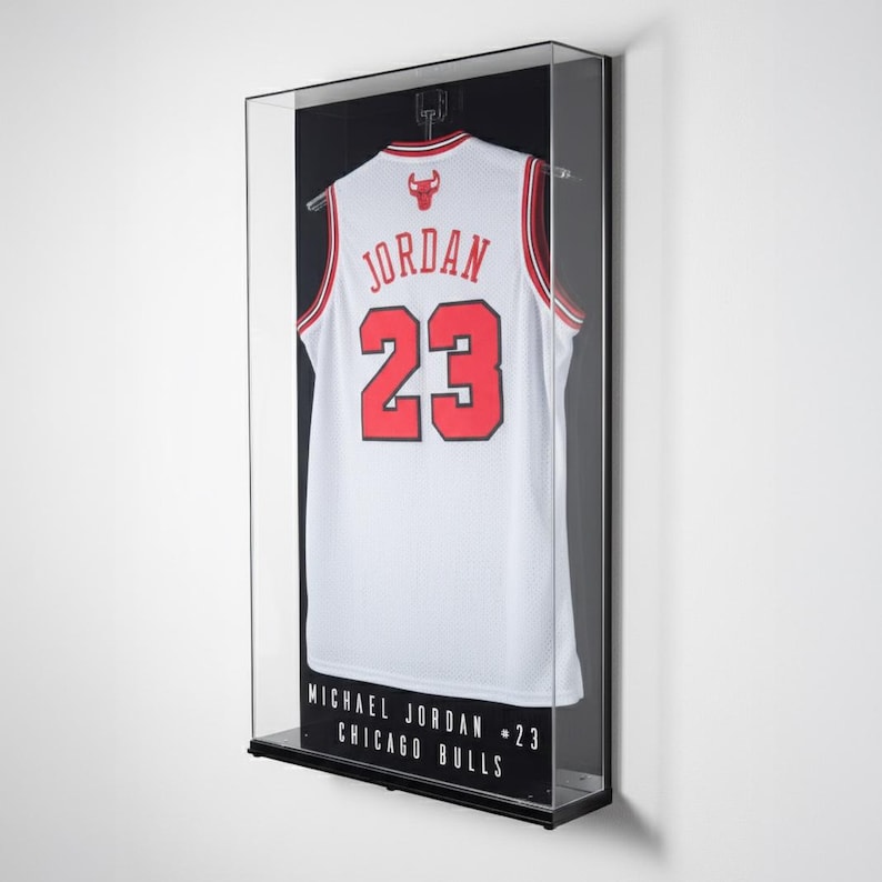 Puede incluir: Una camiseta de baloncesto blanca con detalles rojos y el n&uacute;mero 23, exhibida en una vitrina con marco negro. La camiseta tiene la palabra "JORDAN" en el pecho. La vitrina incluye el texto "MICHAEL JORDAN - 23 CHICAGO BULLS".