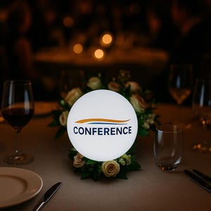 May include: A white, illuminated sphere with the word "CONFERENCE" in blue and a gold and blue design. The sphere sits on a table with a floral arrangement and is surrounded by glasses and tableware, suggesting a conference or event setting.