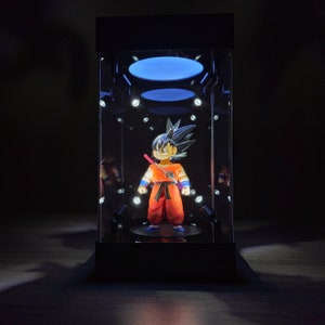 May include: A Dragon Ball Z action figure of Goku in an orange gi, black hair, and blue boots, displayed in a black, illuminated display case. The case has a clear front and a black base, with spotlights and a blue light.