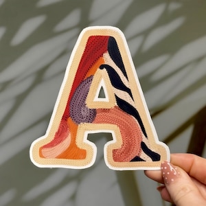 Puede incluir: Un parche decorativo con la forma de la letra "A". El parche presenta un borde color crema y está relleno con una variedad de colores, incluyendo rojo, naranja, morado y negro. El diseño tiene una apariencia texturizada y cosida.
