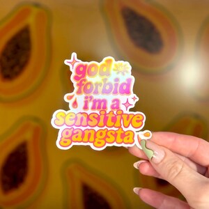 K&ouml;nnte beinhalten: Ein farbenfroher Aufkleber mit dem Text "god forbid i'm a sensitive gangsta" in einer verspielten Schriftart. Der Aufkleber hat einen wei&szlig;en Rand und wird vor einem Hintergrund aus Papaya-Scheiben gehalten.