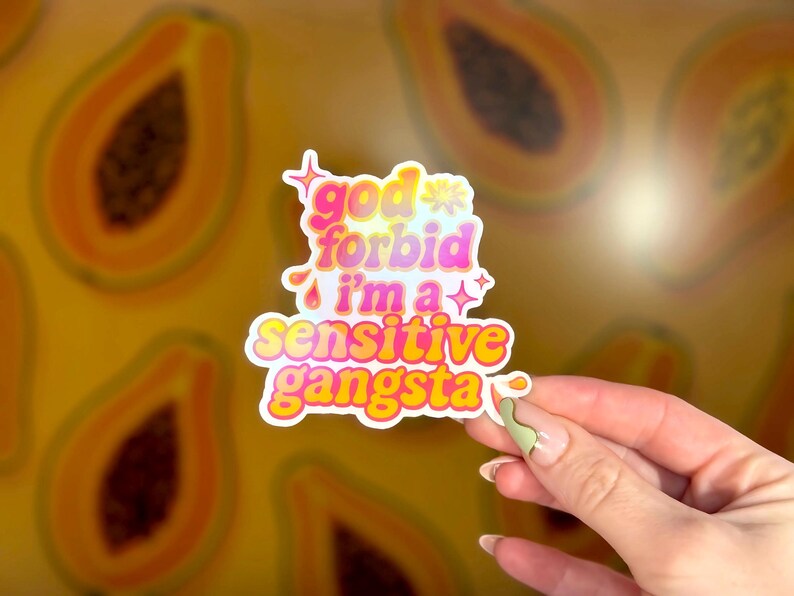 K&ouml;nnte beinhalten: Ein farbenfroher Aufkleber mit dem Text "god forbid i'm a sensitive gangsta" in einer verspielten Schriftart. Der Aufkleber hat einen wei&szlig;en Rand und wird vor einem Hintergrund aus Papaya-Scheiben gehalten.