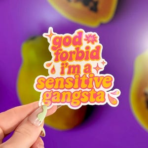 K&ouml;nnte beinhalten: Ein Aufkleber mit dem Text "god forbid i'm a sensitive gangsta" in Pink und Gelb, mit wei&szlig;em Rand. Der Aufkleber wird vor einem lila Hintergrund mit Papaya-Scheiben gehalten.