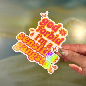 K&ouml;nnte beinhalten: Ein holografischer Aufkleber mit dem Text "god forbid i'm a sensitive gangsta" in Orange und Pink. Der Aufkleber hat einen wei&szlig;en Rand und wird von einer Hand gehalten.