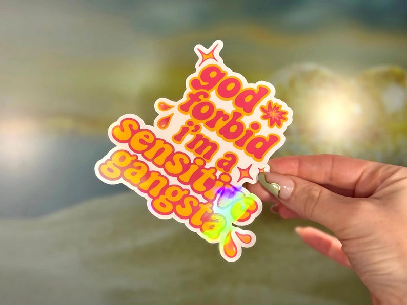 K&ouml;nnte beinhalten: Ein holografischer Aufkleber mit dem Text "god forbid i'm a sensitive gangsta" in Orange und Pink. Der Aufkleber hat einen wei&szlig;en Rand und wird von einer Hand gehalten.