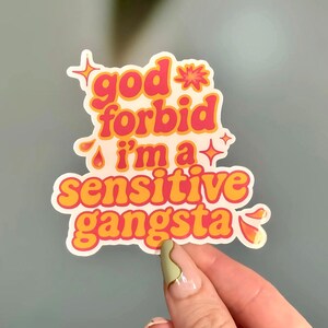 K&ouml;nnte beinhalten: Ein wei&szlig;er Aufkleber mit dem Text "god forbid i'm a sensitive gangsta" in orangefarbenen und rosa Buchstaben. Der Aufkleber hat eine Retro-&Auml;sthetik mit Sternen und Glitzer.