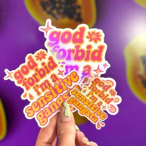 K&ouml;nnte beinhalten: Eine Sammlung von Aufklebern mit dem Text "god forbid I'm a sensitive gangsta" in einem Retro-Schriftstil. Die Aufkleber sind in Pink, Orange und Gelb gehalten und haben einen holografischen Effekt. Der Hintergrund ist lila mit Papaya-Scheiben.