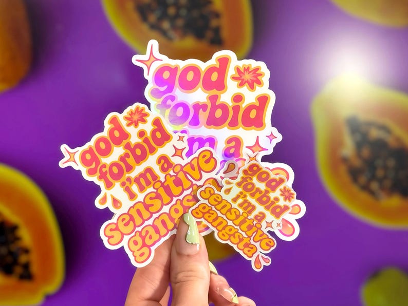 K&ouml;nnte beinhalten: Eine Sammlung von Aufklebern mit dem Text "god forbid I'm a sensitive gangsta" in einem Retro-Schriftstil. Die Aufkleber sind in Pink, Orange und Gelb gehalten und haben einen holografischen Effekt. Der Hintergrund ist lila mit Papaya-Scheiben.