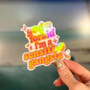 K&ouml;nnte beinhalten: Ein holografischer Aufkleber mit dem Text "god forbid I'm a sensitive gangsta" in einer bunten Schriftart im Blasenstil. Der Aufkleber hat einen wei&szlig;en Rand und wird vor einem verschwommenen Hintergrund einer Strandszene gehalten.