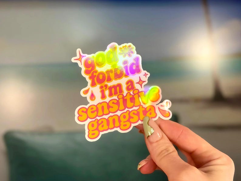 K&ouml;nnte beinhalten: Ein holografischer Aufkleber mit dem Text "god forbid I'm a sensitive gangsta" in einer bunten Schriftart im Blasenstil. Der Aufkleber hat einen wei&szlig;en Rand und wird vor einem verschwommenen Hintergrund einer Strandszene gehalten.