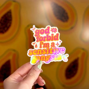 K&ouml;nnte beinhalten: Ein farbenfroher Aufkleber mit dem Text "god forbid i'm a sensitive gangsta" in Pink, Gelb und Lila. Der Aufkleber hat einen wei&szlig;en Rand und ist vor einem Hintergrund mit Papaya-Fr&uuml;chten platziert.