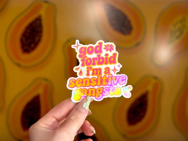 K&ouml;nnte beinhalten: Ein farbenfroher Aufkleber mit dem Text "god forbid i'm a sensitive gangsta" in Pink, Gelb und Lila. Der Aufkleber hat einen wei&szlig;en Rand und ist vor einem Hintergrund mit Papaya-Fr&uuml;chten platziert.