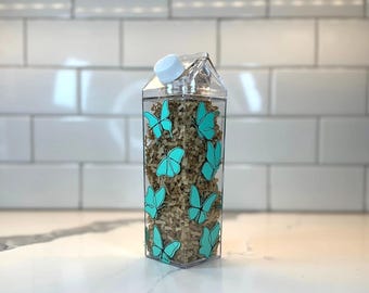 Botella de agua reutilizable de cartón de leche con diseño de mariposas en azul verdoso.