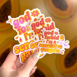 K&ouml;nnte beinhalten: Eine Sammlung von Aufklebern mit dem Text "god forbid i'm a sensitive gangsta" in rosa und gelber Schrift. Die Aufkleber haben einen wei&szlig;en Rand und Stern- und Tropfendesigns. Der Hintergrund ist ein verschwommenes Bild von Papaya.