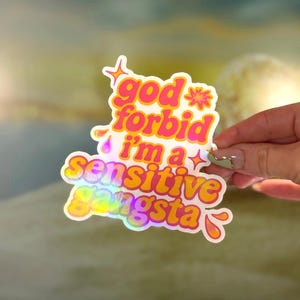 K&ouml;nnte beinhalten: Ein holografischer Aufkleber mit dem Text "god forbid i'm a sensitive gangsta" in einer verspielten, farbenfrohen Schriftart. Der Aufkleber hat einen wei&szlig;en Rand und wird von einer Hand gehalten.