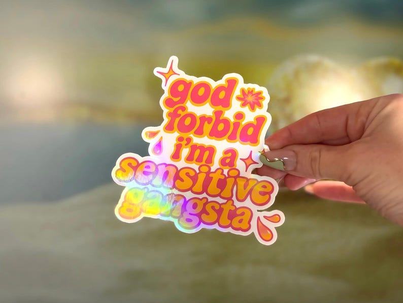 K&ouml;nnte beinhalten: Ein holografischer Aufkleber mit dem Text "god forbid i'm a sensitive gangsta" in einer verspielten, farbenfrohen Schriftart. Der Aufkleber hat einen wei&szlig;en Rand und wird von einer Hand gehalten.