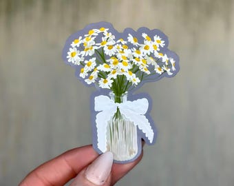 Puede incluir: Una pegatina con un ramo de margaritas en un tarro de cristal transparente, adornado con una cinta blanca. Las margaritas tienen pétalos blancos y centros amarillos. La pegatina tiene un contorno morado claro.