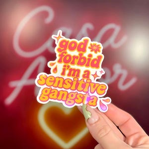 K&ouml;nnte beinhalten: Ein farbenfroher Aufkleber mit dem Text "god forbid i'm a sensitive gangsta" in Pink und Gelb. Das Retro-Design enth&auml;lt Sterne und Blumen. Der Hintergrund ist ein verschwommenes Rot mit einem Neonherz.