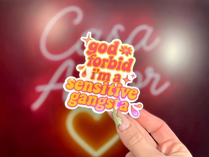 K&ouml;nnte beinhalten: Ein farbenfroher Aufkleber mit dem Text "god forbid i'm a sensitive gangsta" in Pink und Gelb. Das Retro-Design enth&auml;lt Sterne und Blumen. Der Hintergrund ist ein verschwommenes Rot mit einem Neonherz.