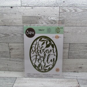 Puede incluir: Un paquete de troqueles Sizzix Thinlits. El troquelado es de forma ovalada con la frase "the season to be jolly" en escritura cursiva, rodeada de hojas y acebo. El paquete también incluye el texto "2 DIES 2 MATRICES 2 TROQUELES 2 FUSTELLE".