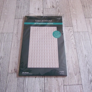 Puede incluir: Una carpeta de relieve 3D Spellbinders en un paquete de plástico transparente. La carpeta presenta un diseño de mosaico de azulejos con un patrón geométrico repetitivo. El paquete también incluye texto que dice "Create 3D Textured A2, Slimline, 5x7" Cards & More!"