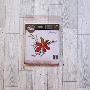May include: A Sizzix Thinlits die set featuring a red poinsettia design with green leaves and sprigs. The package includes the text "Tim Holtz" and "8 Dies". The background is a light wood grain.
