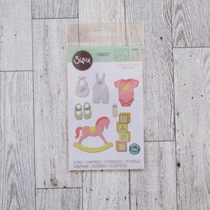 May include: A die-cut set featuring baby-themed shapes, including a bib, overalls, a onesie, baby shoes, a bottle, a rocking horse, and alphabet blocks. The set is in pastel colors and includes the text "Sizzix" and "THINLITS".
