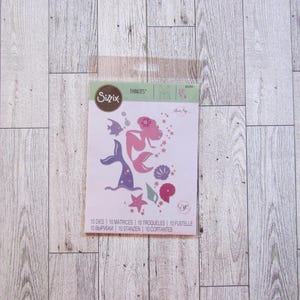 Puede incluir: Un paquete de troqueles Sizzix Thinlits con temática de sirenas. Las formas troqueladas incluyen una sirena, peces, estrellas de mar y conchas en rosa, morado y azul. El paquete incluye el texto "10 DIES 10 MATRICES 10 TROQUELES 10 FUSTELLE".