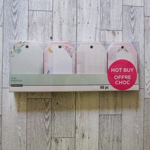 May include: A package of 60 gift tags in a clear plastic container. The tags have floral designs and a pink and white color scheme. The package includes the text "TAGS ETIQUETTES" and a "HOT BUY OFFRE CHOC" sticker.