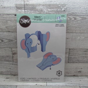 May include: A package of Sizzix Thinlits Fold-A-Long dies. The package features a blue elephant design with pink ears. The package includes 6 dies, 6 matrices, 6 troqueles, 6 fustelle, 6 vyrubki, 6 stanzen, and 6 cortantes.