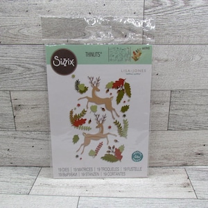May include: A Sizzix Thinlits die set designed by Lisa Jones. The package shows a design with two deer, leaves, and floral elements in brown, red, and green. The set contains 19 dies for crafting projects.