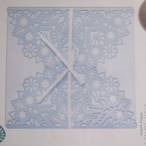 May include: A pale blue, die-cut paper craft featuring a snowflake and floral pattern. The paper is folded and secured with a white ribbon. The design incorporates small, sparkling embellishments.
