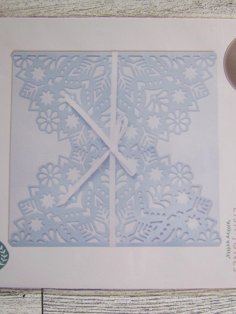 May include: A pale blue, die-cut paper craft featuring a snowflake and floral pattern. The paper is folded and secured with a white ribbon. The design incorporates small, sparkling embellishments.