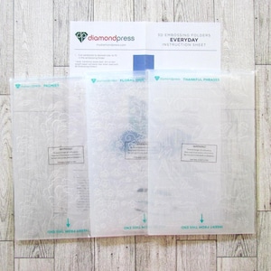 May include: Three clear plastic embossing folders with floral and phrase designs, labeled "Peonies", "Floral Sprays", and "Thankful Phrases". An instruction sheet with the Diamond Press logo is on top. The folders are on a light wood grain surface.