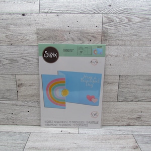 May include: A Sizzix Thinlits die set with a rainbow and sun design. The package includes 10 dies for creating cards with the text "Have a Beautiful Day". The card design includes a rainbow, sun, and hearts.