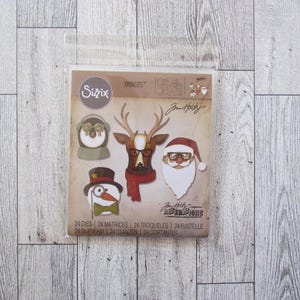 May include: A pack of Sizzix Thinlits dies with Christmas-themed characters. The designs include a penguin, reindeer, snowman, and Santa Claus, all wearing glasses. The package includes 24 dies and 24 matrices.