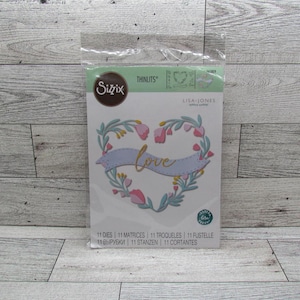 Puede incluir: Un paquete de troqueles Sizzix Thinlits con un diseño floral en forma de corazón y la palabra "love" en una pancarta. El diseño incluye flores rosas, hojas verdes y una pancarta azul claro. Contiene 11 troqueles.